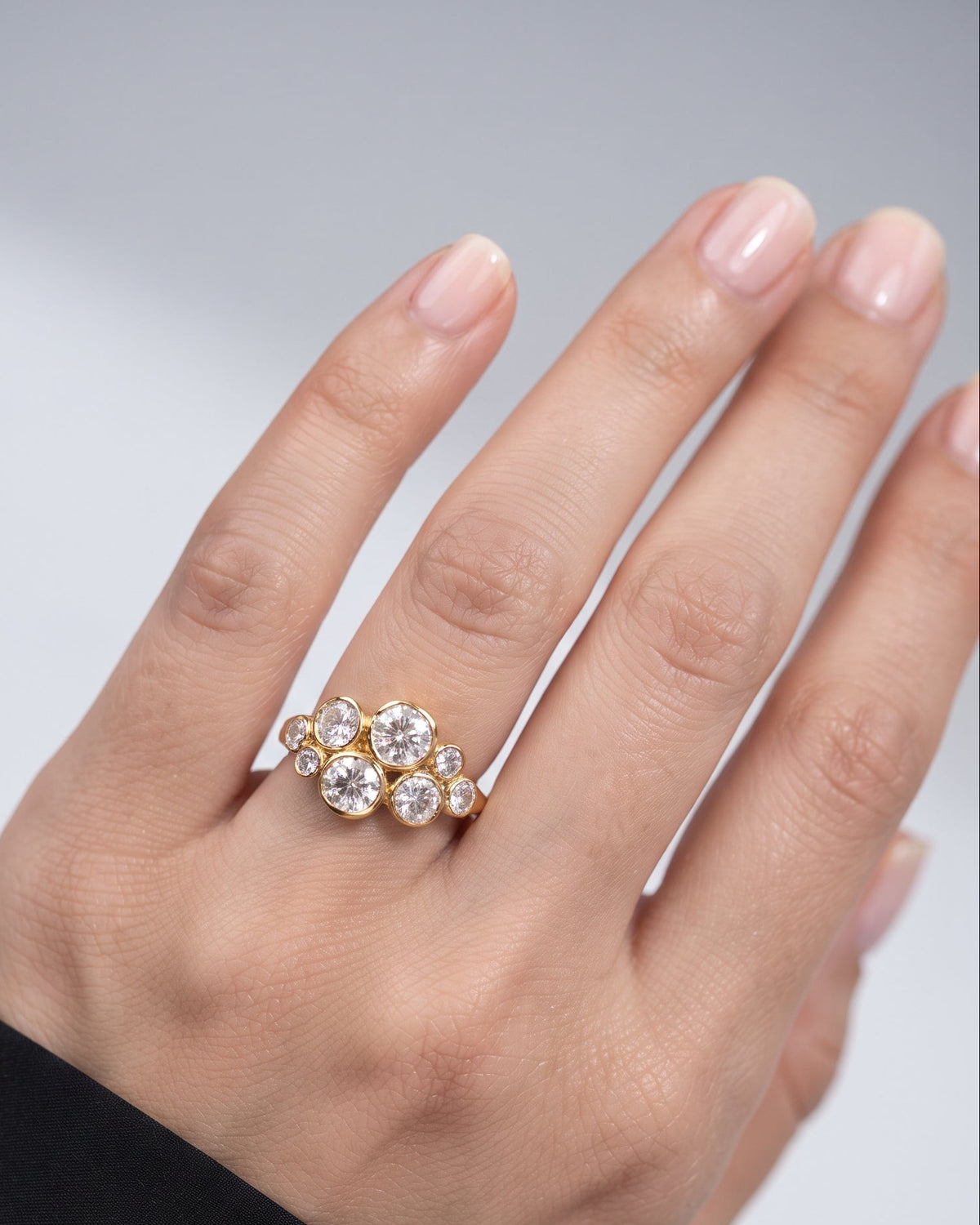 A close-up of a hand wearing a gold ring with a cluster of clear round-cut gemstones, set against a soft light gray background.