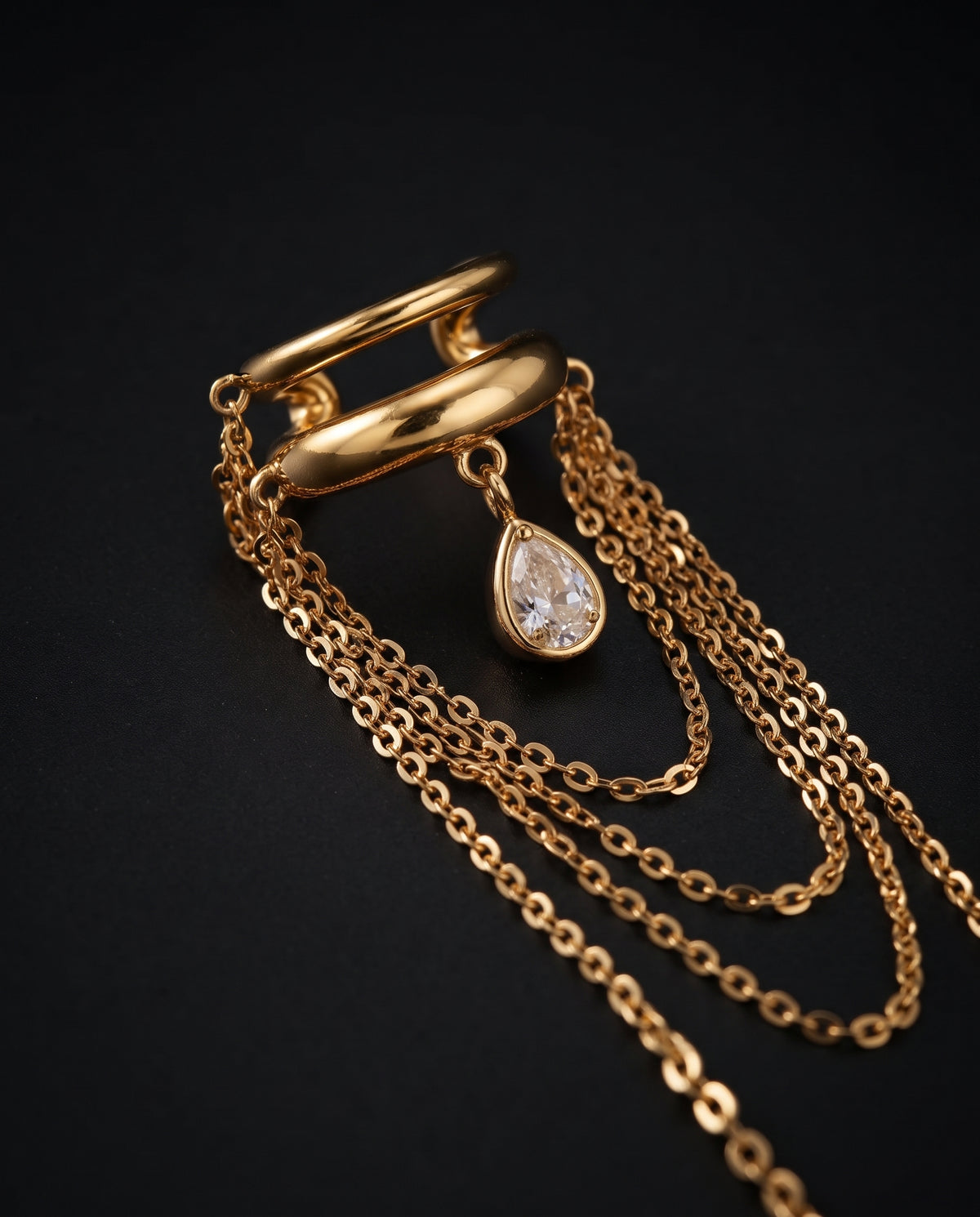 A gold-toned layered chain necklace with a teardrop-shaped clear gemstone pendant, displayed on a dark black background.