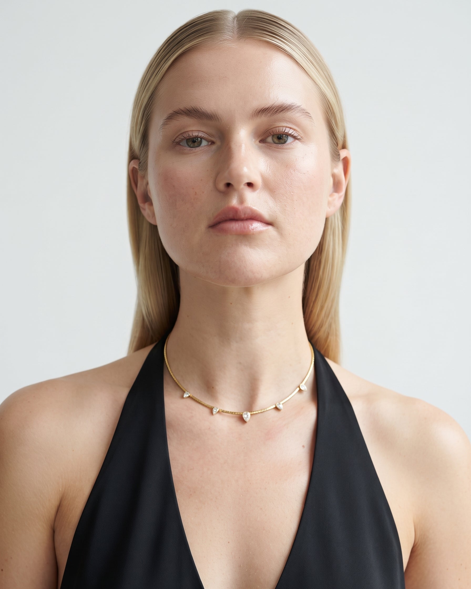 A blonde woman wearing a delicate gold chain necklace with small white bead accents, styled with a black halter top against a light gray background.
