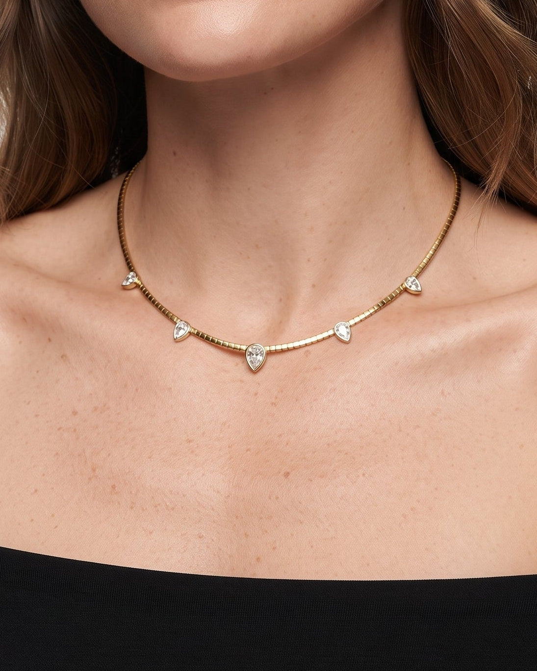 A woman wearing a delicate gold chain necklace with small gold and clear crystal accents, styled with a black off-the-shoulder top against a clean white background.