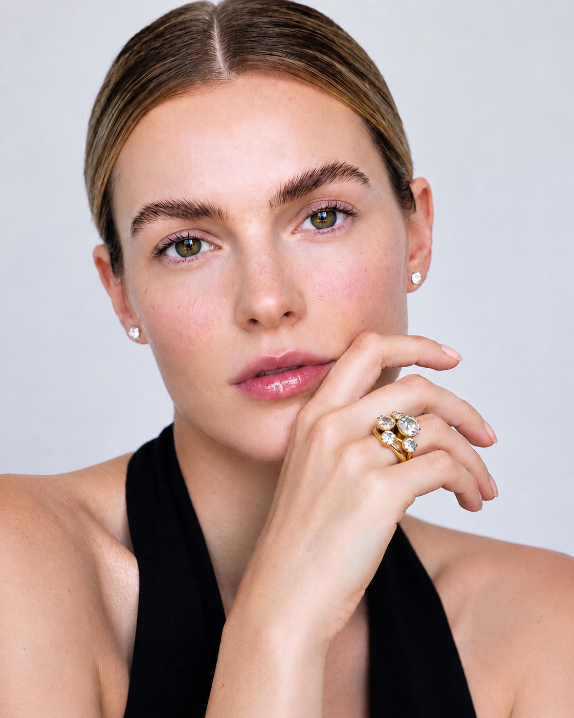 A close-up portrait of a woman wearing a gold ring with clear gemstones on her finger, set against a light gray background.