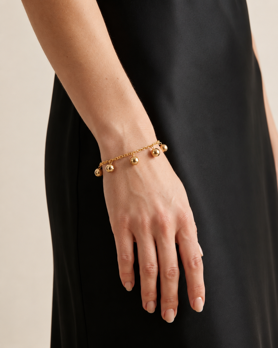 A close-up of a person's hand and wrist wearing a delicate gold chain bracelet with small spherical charms, styled with a black dress against a light beige background.