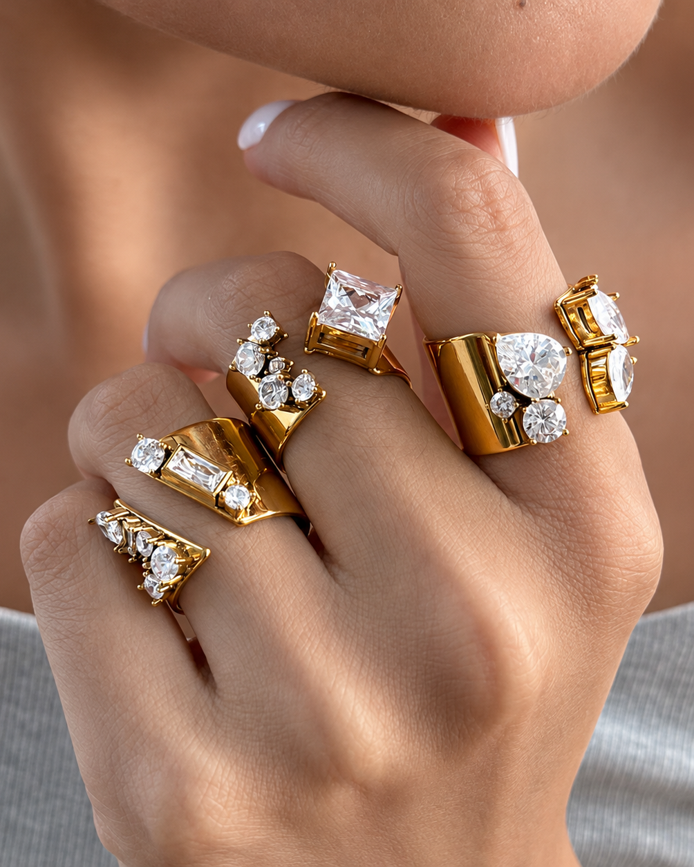 A close-up of a hand wearing multiple gold rings with large clear gemstones, shown on a person's fingers against a soft neutral background.