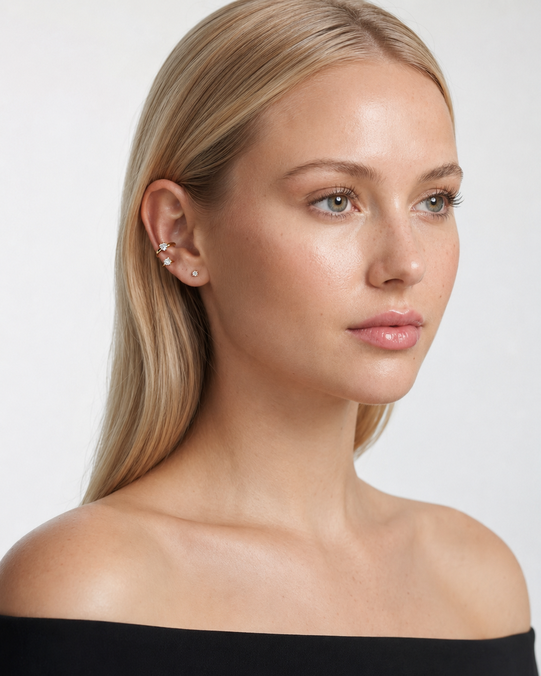 A close-up portrait of a blonde woman wearing small stud earrings and a stack of ear cuffs against a plain light gray background.