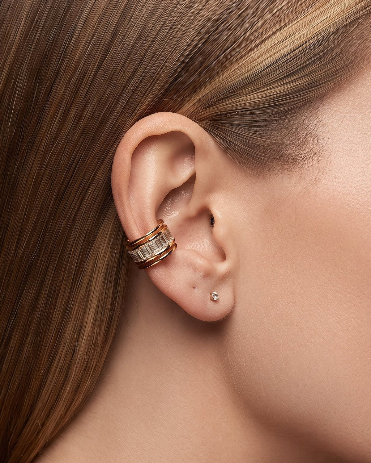 A close-up of a woman's ear wearing a gold ear cuff with clear stones, along with a small stud earring, against a dark background.
