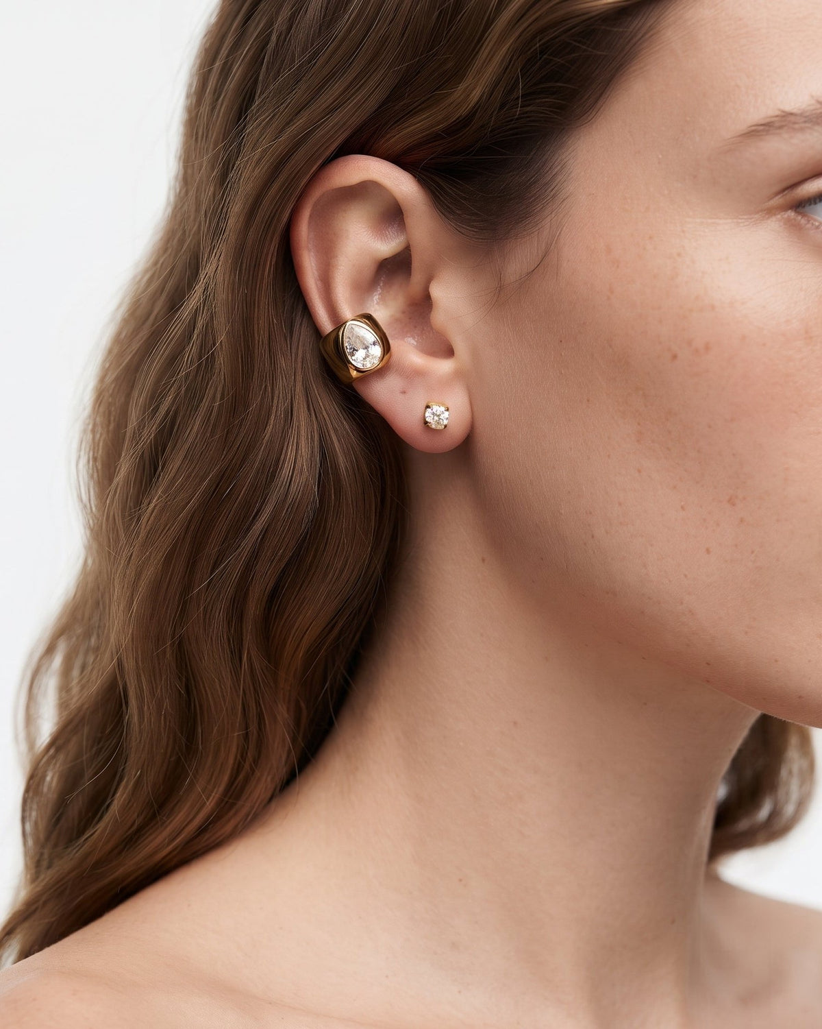 Close-up of a woman's ear wearing a gold post cartilage piercing with a clear oval gemstone and a small silver stud earring, against a light gray background.