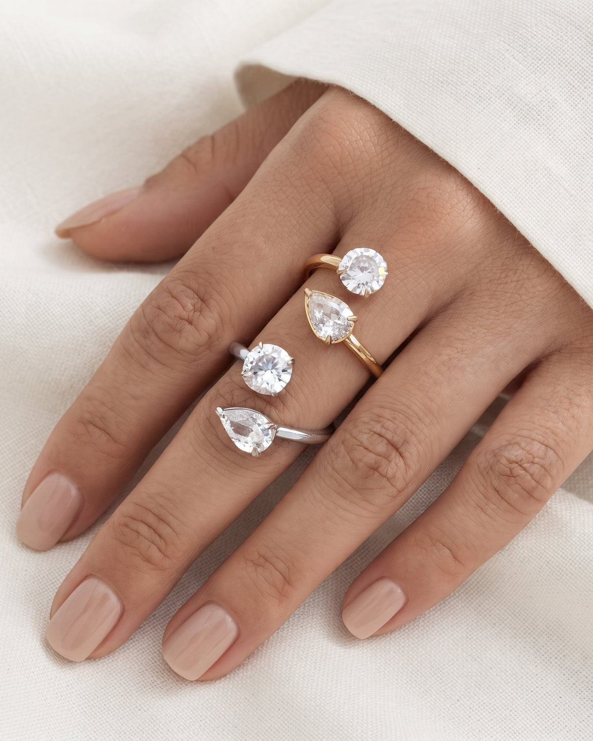 A close-up of a hand wearing four pear-cut gemstone rings in different metal finishes: three silver-toned and one gold-toned. The hand is posed against a soft, draped off-white fabric background.