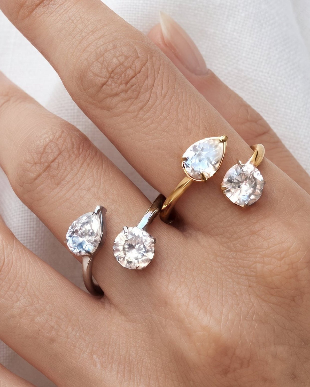 A close-up of a hand wearing four teardrop-shaped gemstone rings in different metal finishes, including silver, yellow gold, and white gold, set against a soft white fabric background.