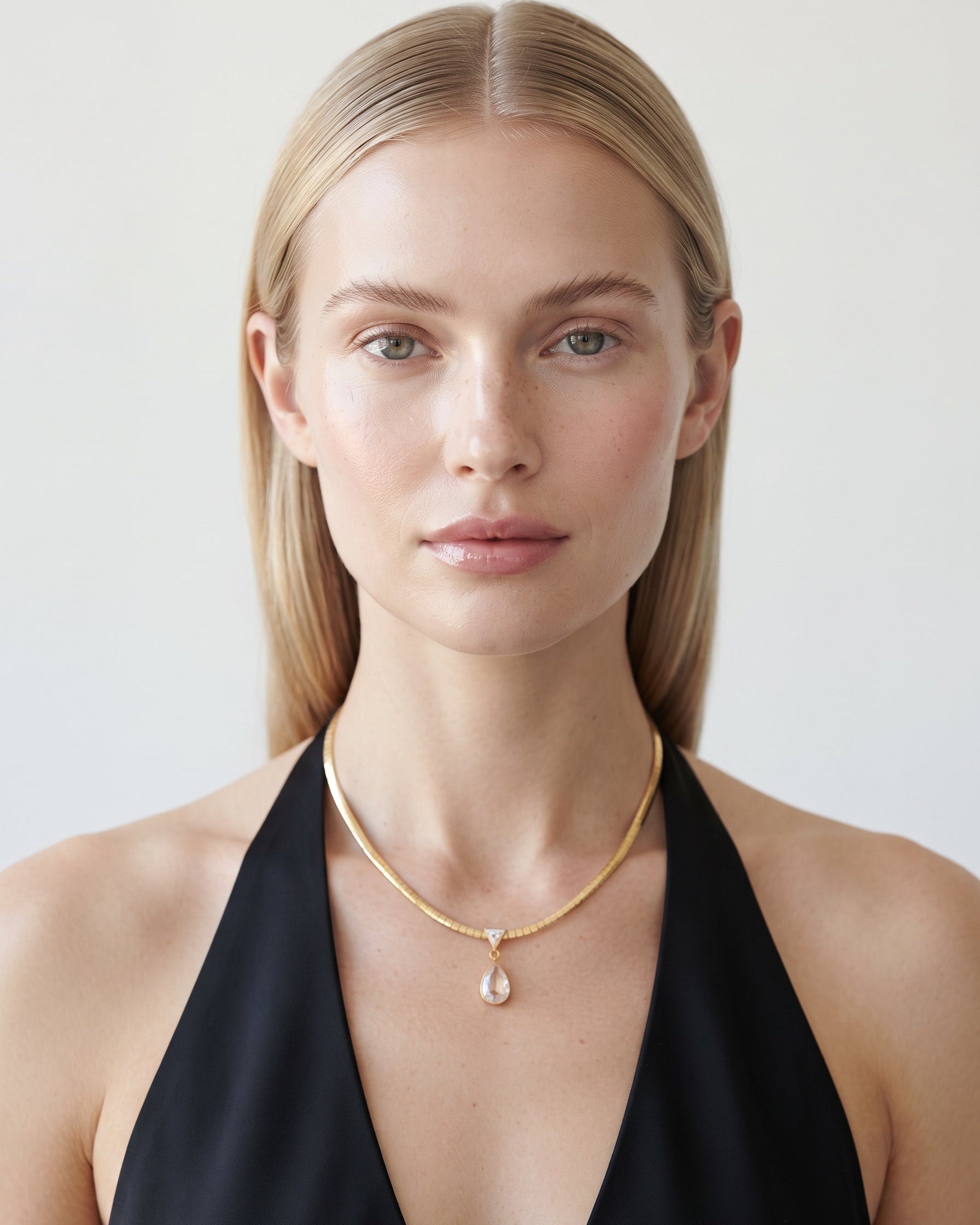 A blonde woman wearing a delicate gold chain necklace with a teardrop-shaped clear gemstone pendant, styled with a black halter top against a light gray background.