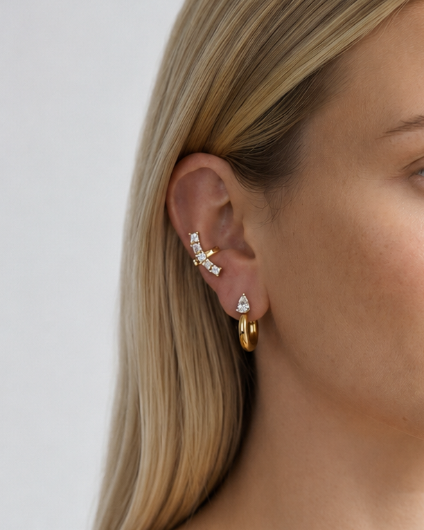A close-up of a blonde woman wearing a gold ear cuff with small clear stones and a gold hoop earring, shown against a light gray background.