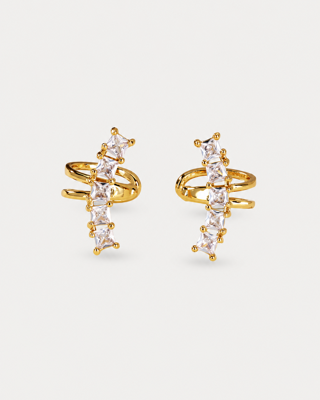A pair of gold wrap-around huggie hoop earrings decorated with a vertical line of clear baguette-cut crystals, set against a light gray background.