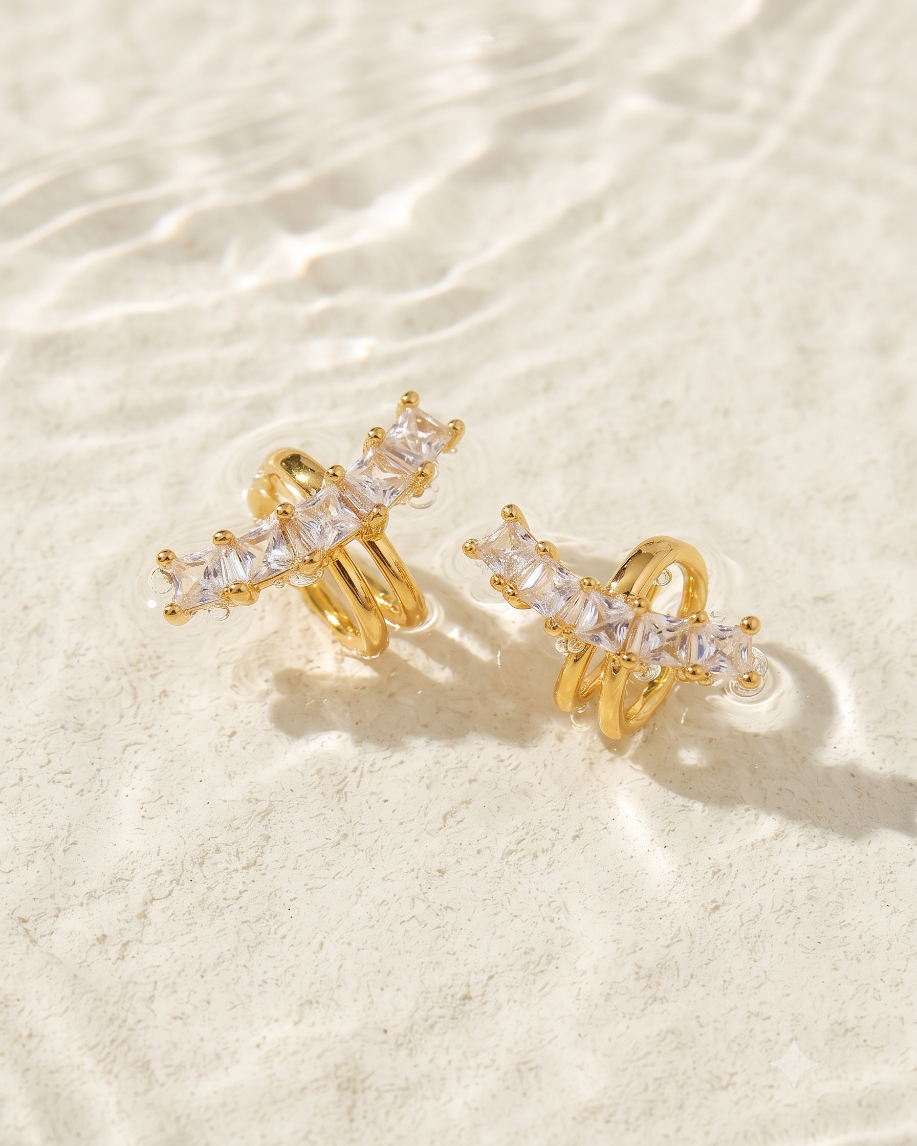 A pair of gold-plated huggie hoop earrings with clear rectangular gemstones, displayed on a textured white surface with soft shadows.