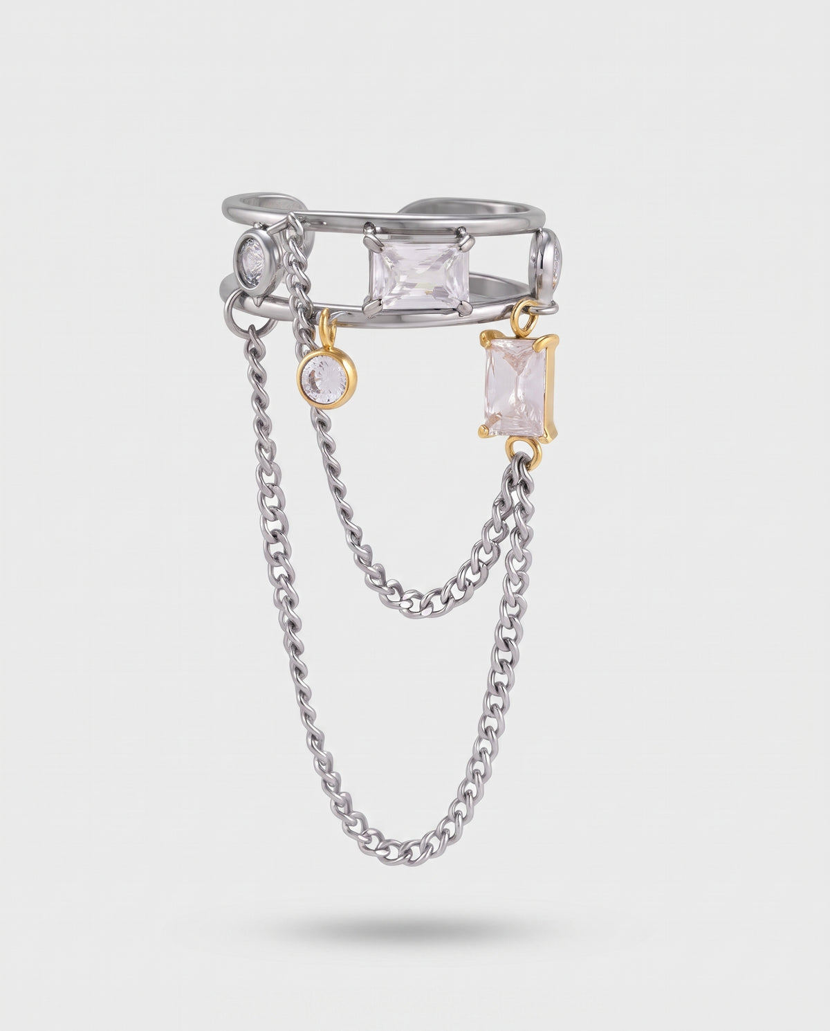 A silver-toned double-layer ring with a top band featuring a clear rectangular gemstone, a dangling clear round gemstone, and a gold-toned dangling rectangular gemstone, with two draped silver chains below, on a light gray background.