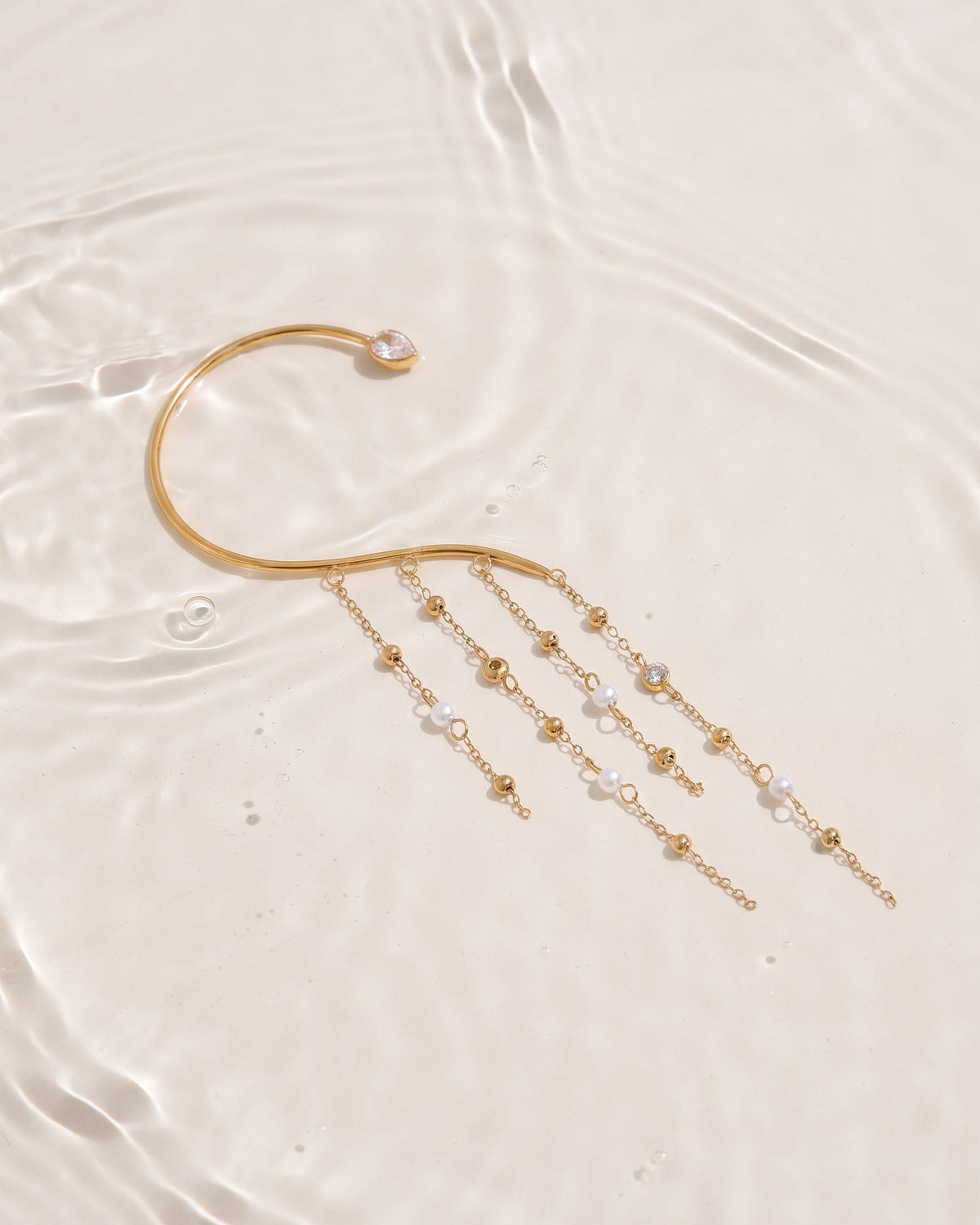 A gold-toned open cuff earring with dangling chains and small clear beads, arranged on a glossy white surface with soft water ripples.