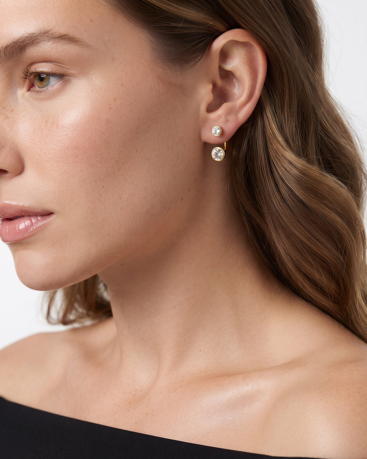 A close-up of a woman's ear and side profile wearing a delicate gold stud earring with a clear round gemstone. The background is a clean, neutral white studio backdrop.