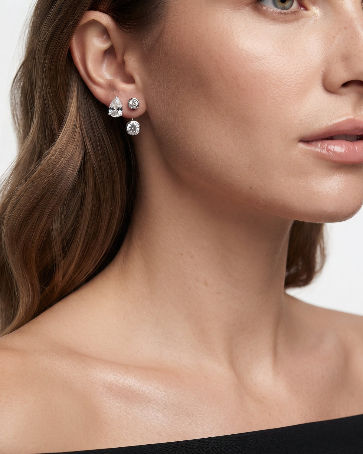 A close-up of a woman wearing a pair of silver stud earrings with clear teardrop and round gemstones, shown against a clean light background.