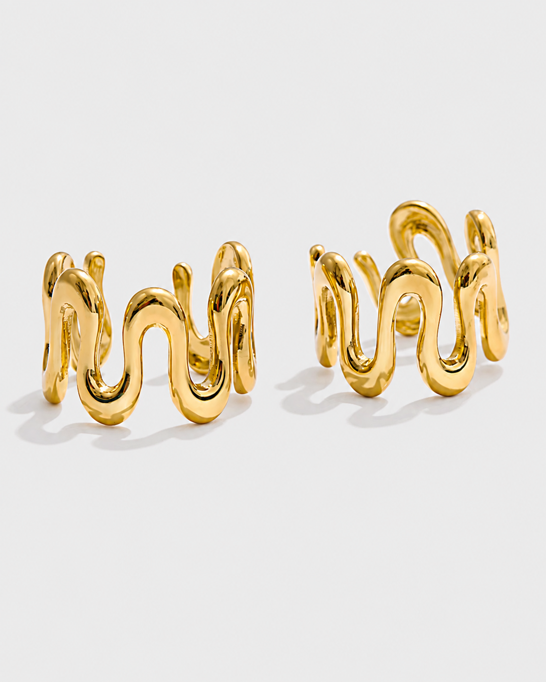 A pair of gold-toned wavy hoop earrings with a thick, sculptural, liquid-like design, displayed on a clean light gray background.