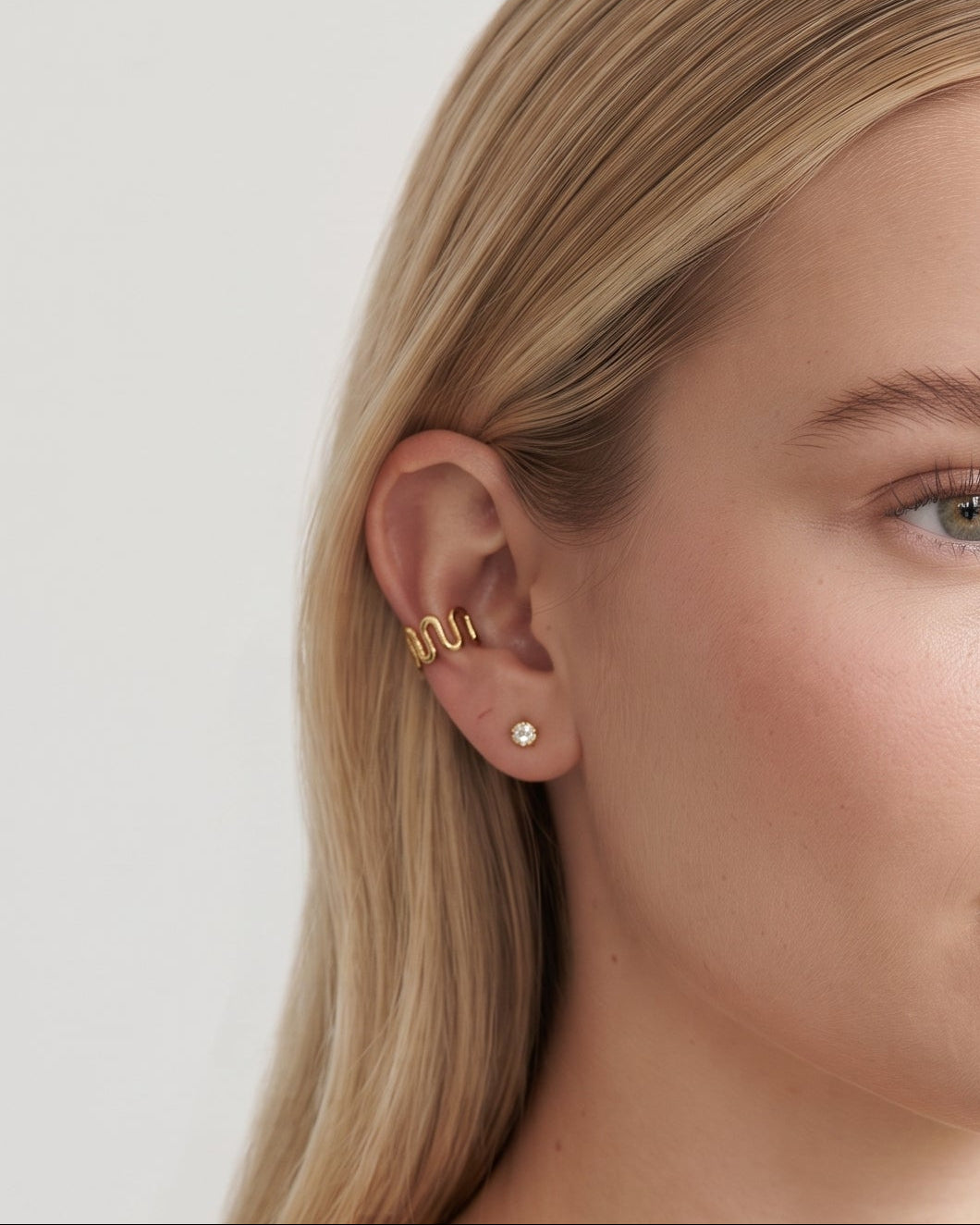 A blonde woman wearing a gold ear cuff with a small pearl stud earring, shown in a close-up portrait against a light gray background.