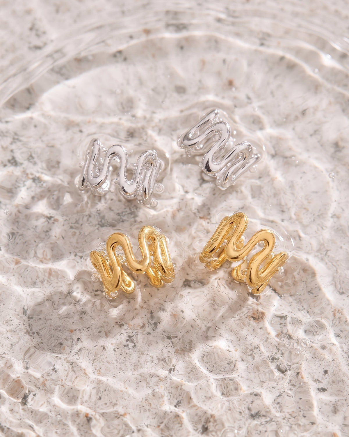 Four wavy stud earrings arranged on a textured stone surface, with two silver-toned earrings and two gold-toned earrings.