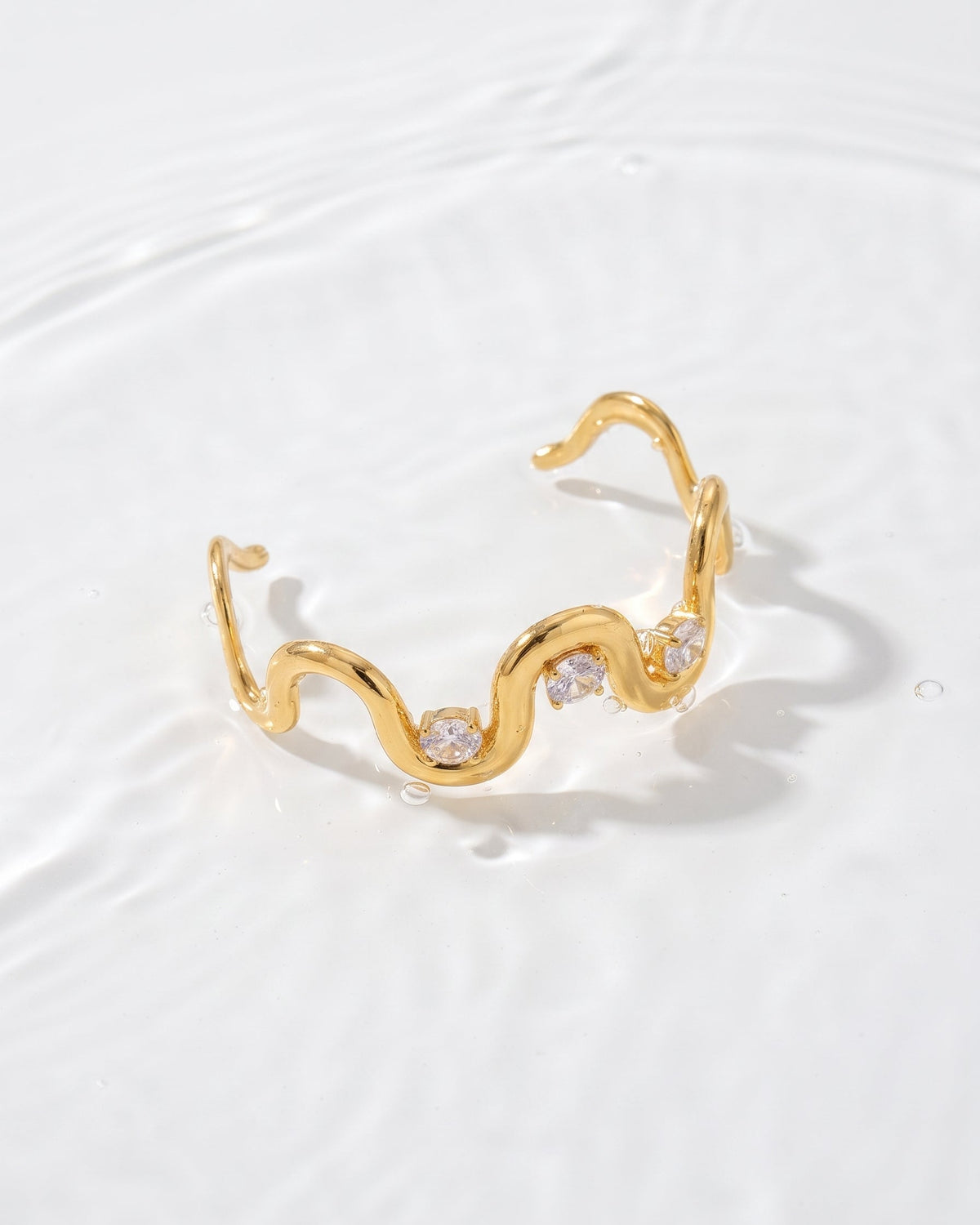 A gold open cuff ring with a wavy, snake-like organic design and three clear round gemstones, resting on a white surface with water ripples.