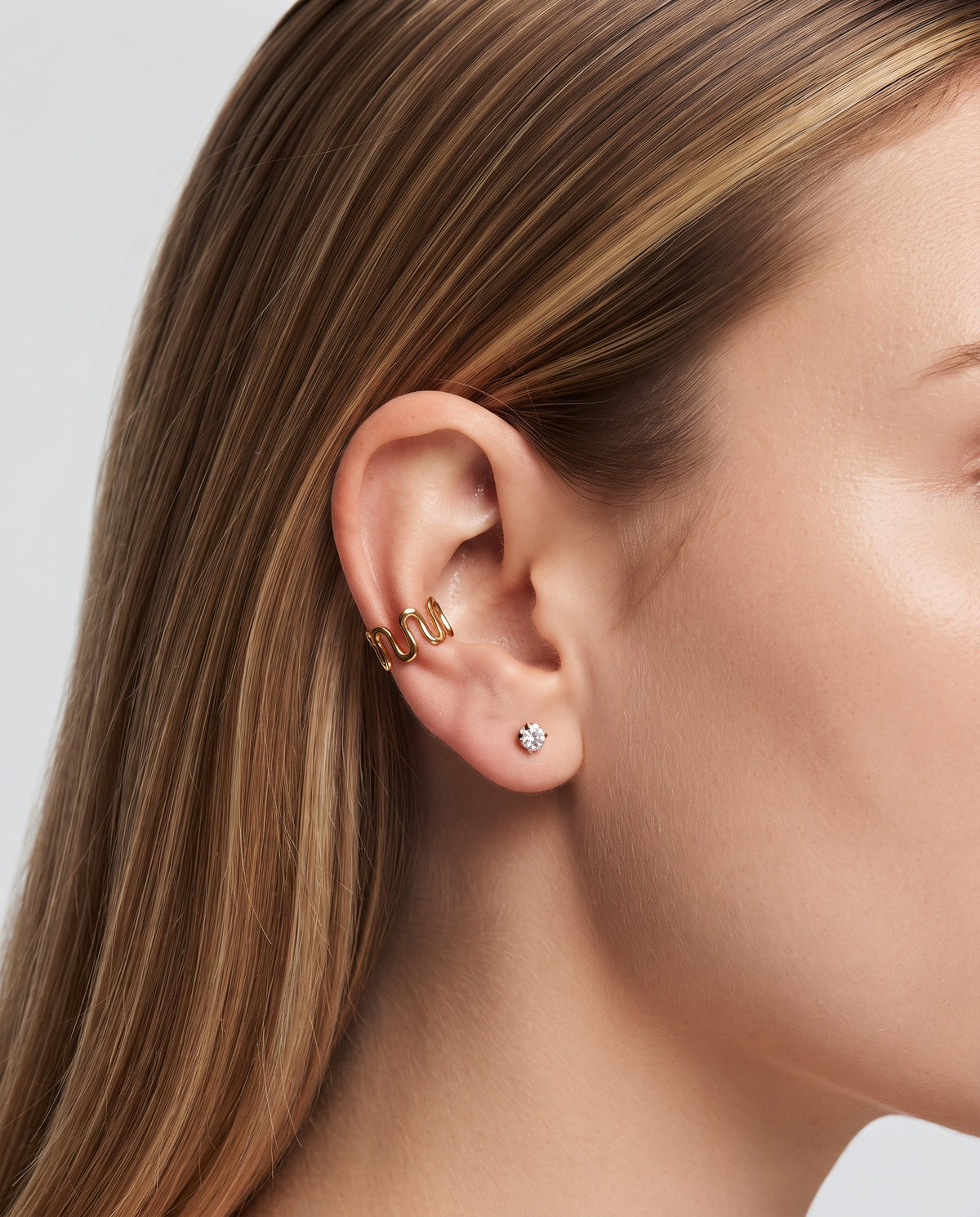 Close-up of a woman's ear wearing two gold ear cuffs and a small silver stud earring, with her hair and neck visible against a light gray background.