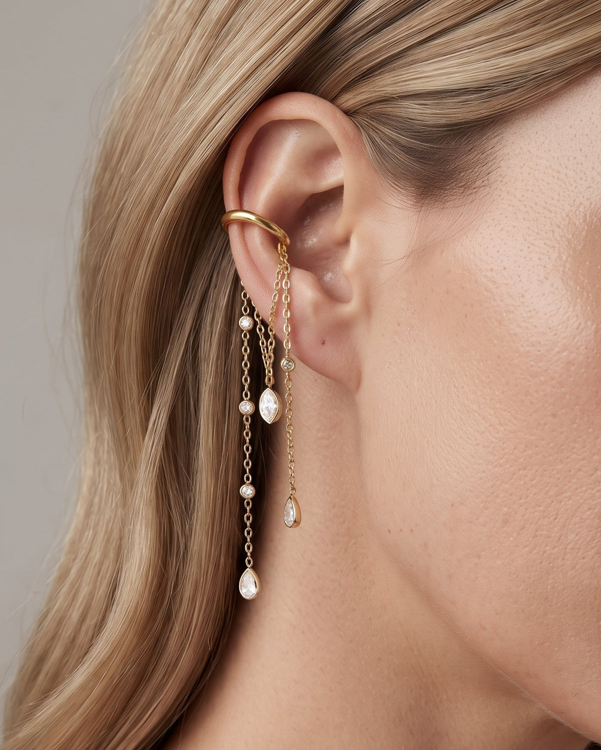A close-up of a model's ear wearing a gold ear cuff with three dangling chain earrings adorned with small white pearls, set against a soft neutral background.