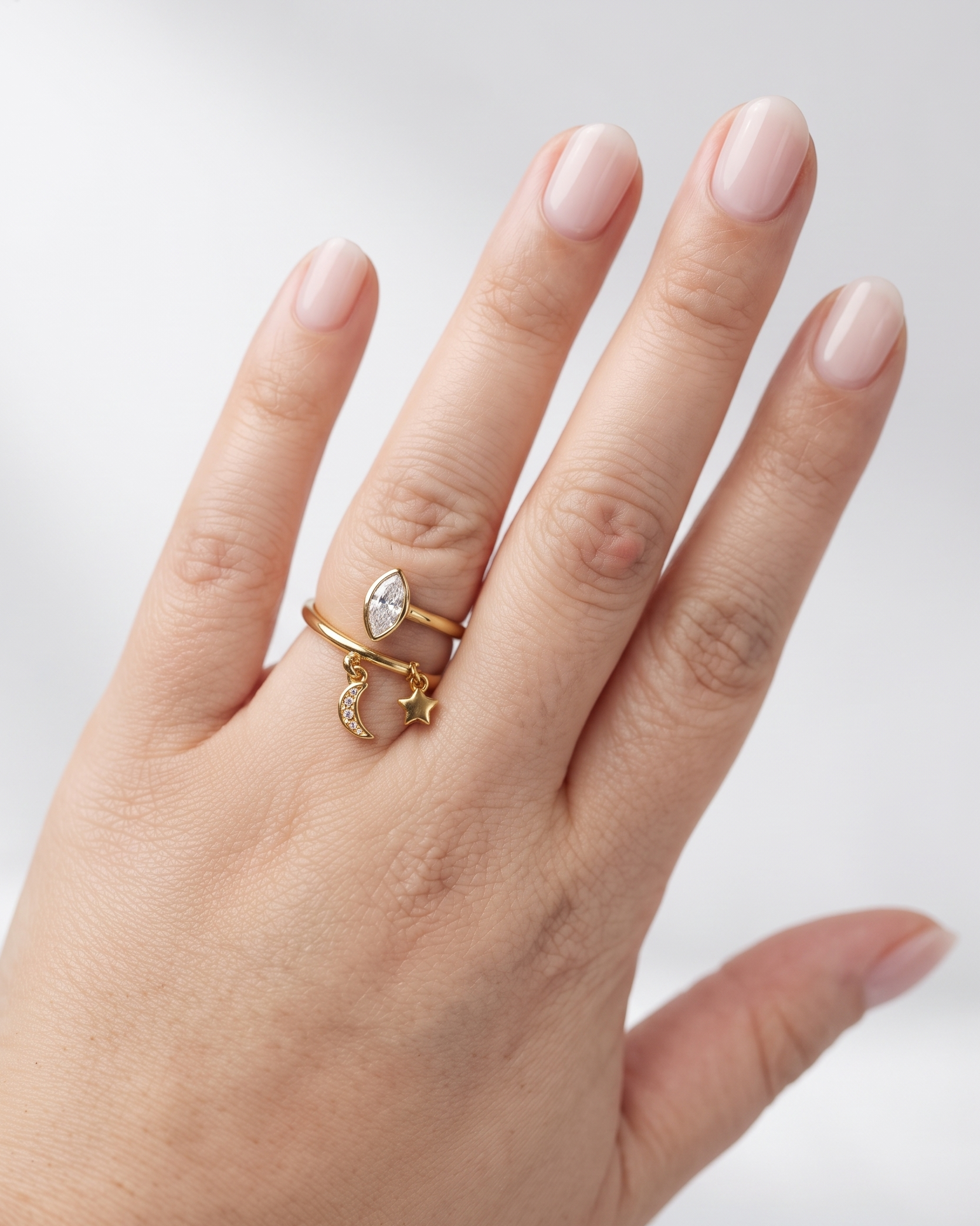 A close-up of a hand wearing two delicate gold rings: one with a small marquise-cut clear gemstone and the other with a small crescent moon charm, set against a soft light gray background.
