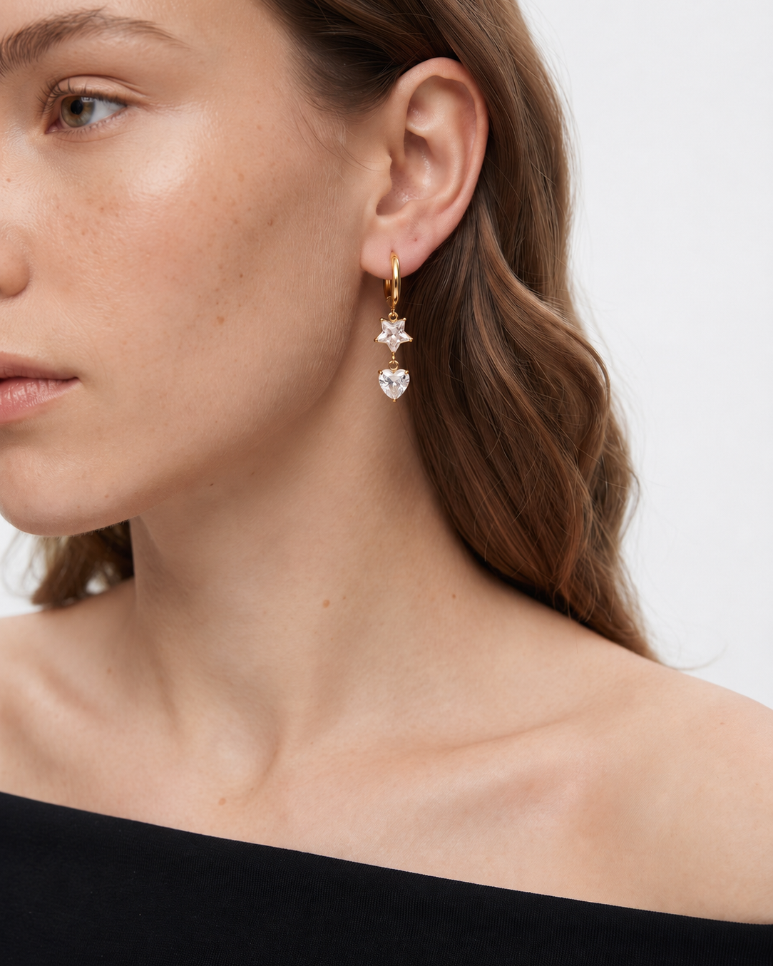 A close-up of a woman wearing a delicate gold hoop earring with two dangling clear heart-shaped crystal charms against a plain light background.