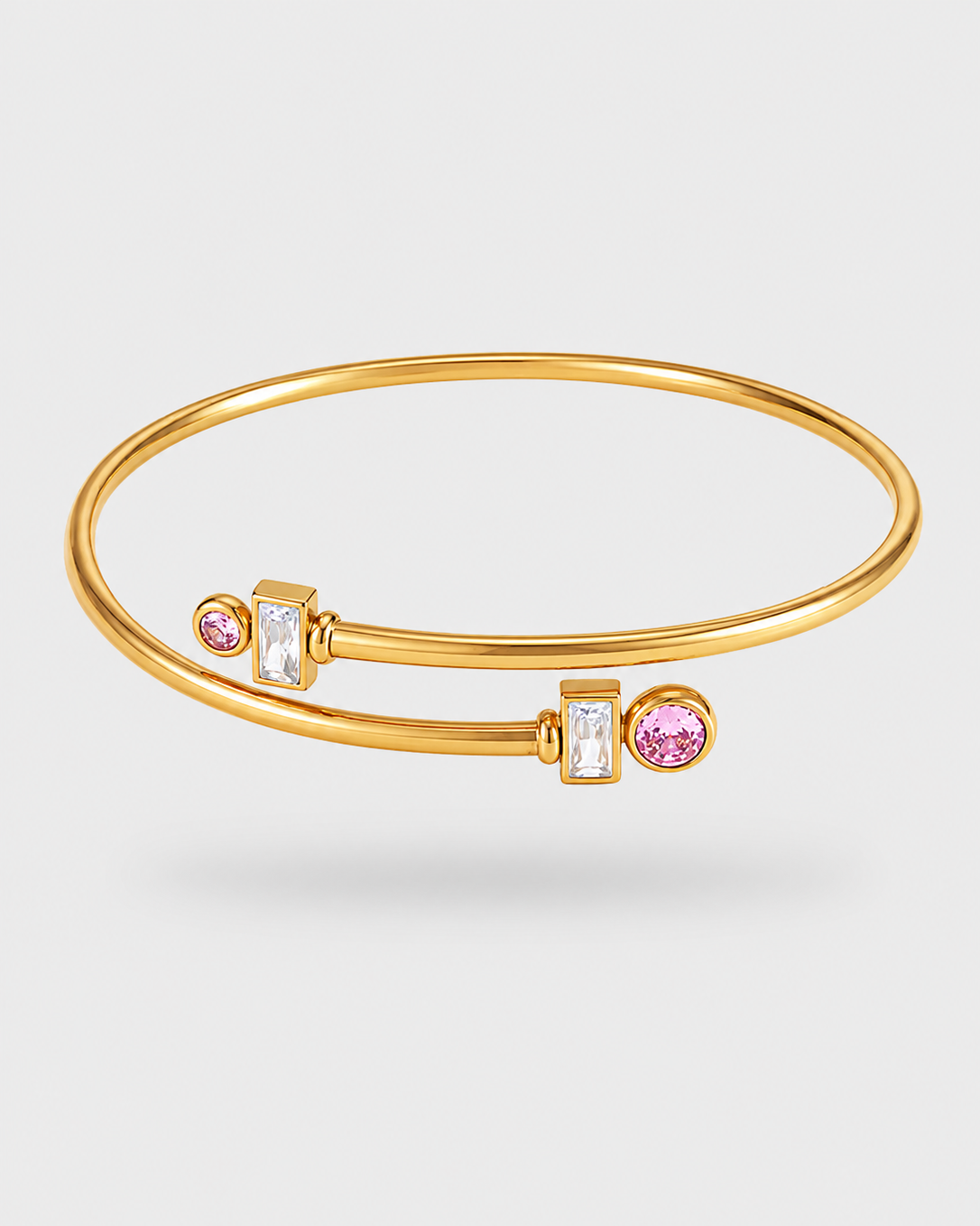 A gold bangle bracelet with two toggle clasp ends, one featuring a round pink gemstone and one featuring a baguette-cut clear gemstone, set against a light gray background.