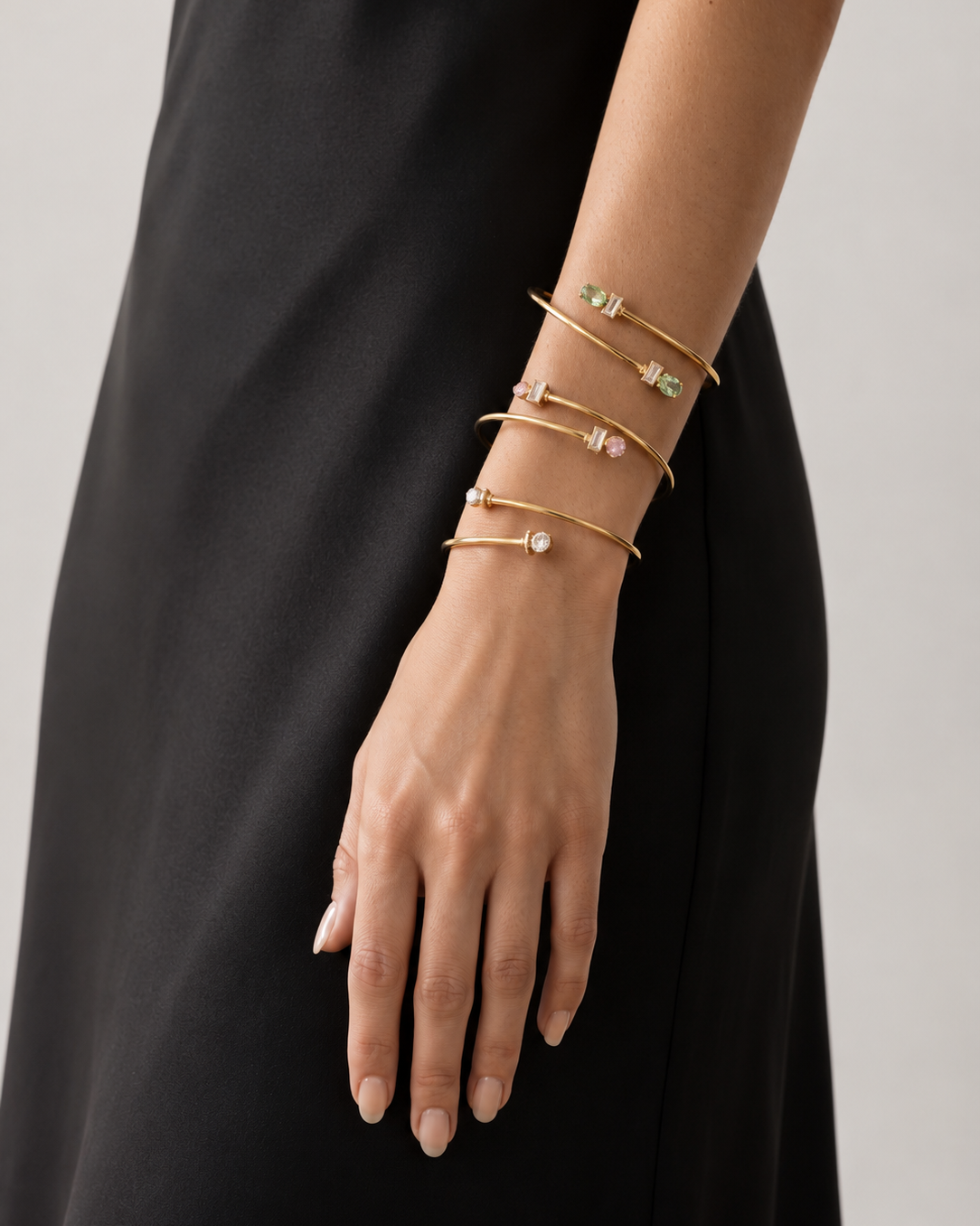 A close-up of a person's wrist and hand wearing five delicate gold bangle bracelets, some adorned with small green and pink gemstones and one with a clear stone, set against a black dress and a light gray background.