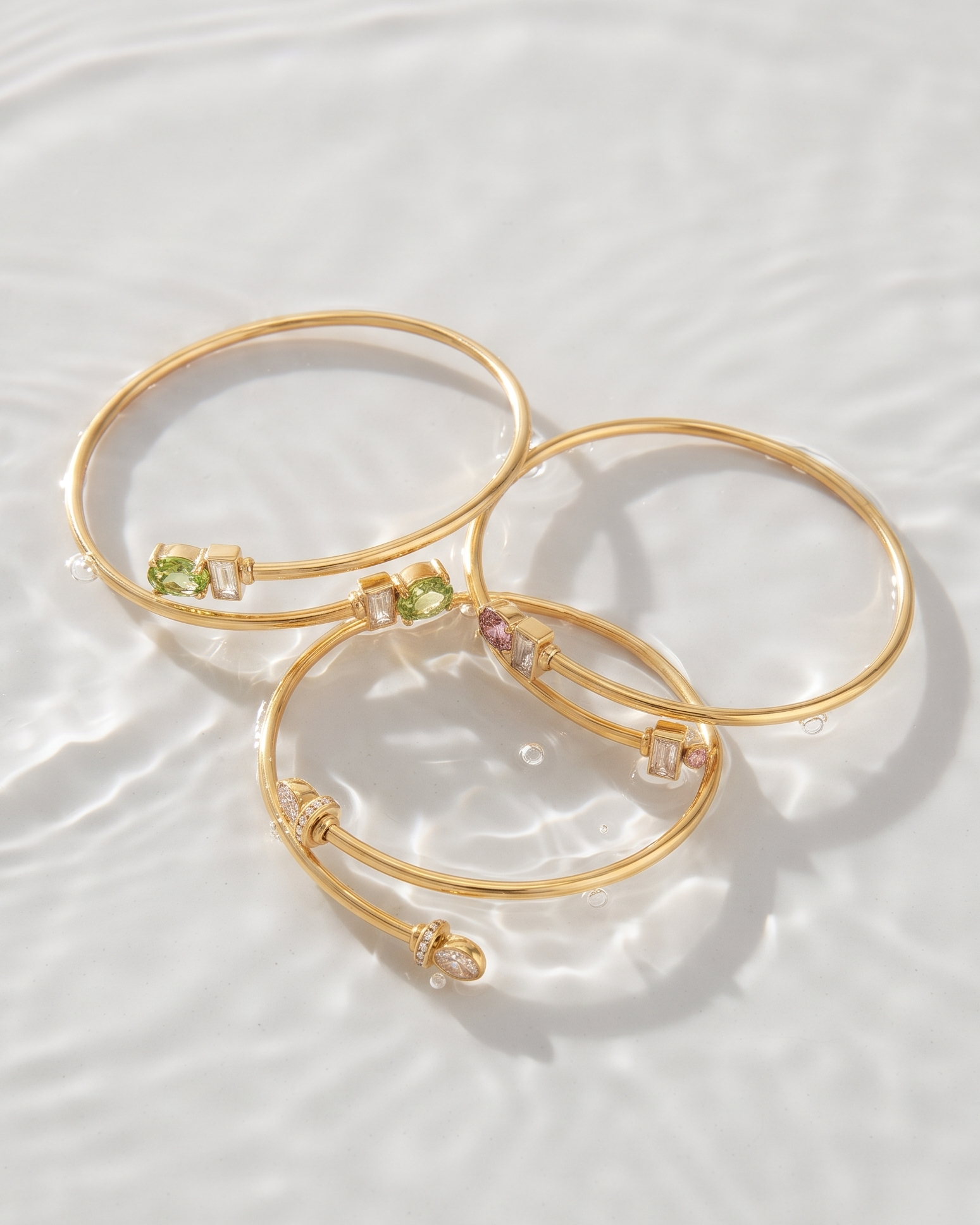 Three gold bangle bracelets with small green and pink gemstone accents, arranged on a white water surface with soft ripples.