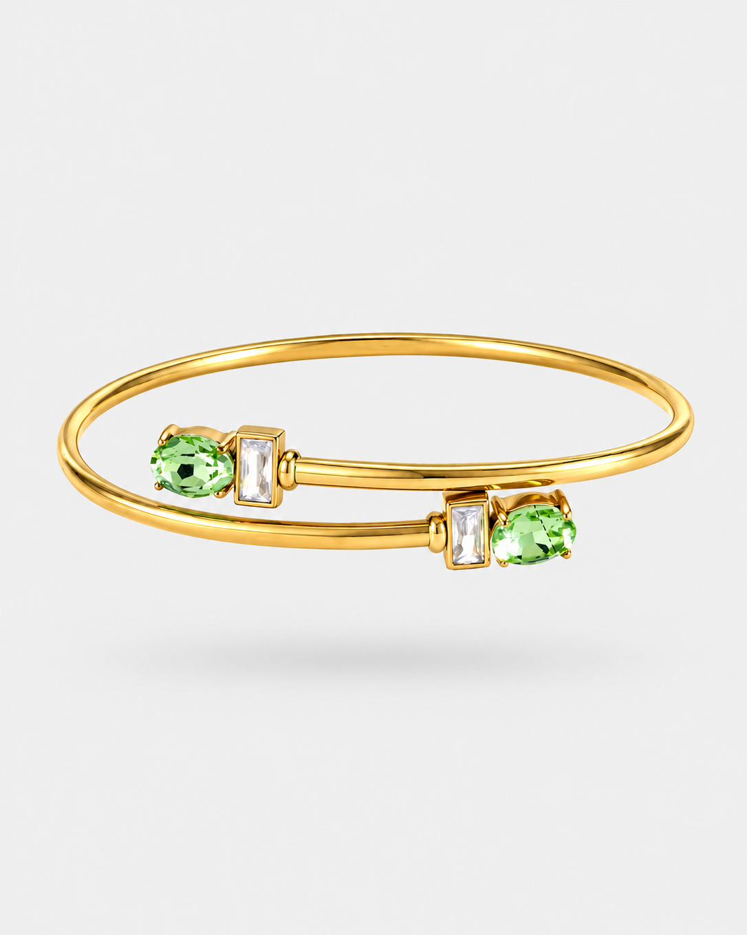 A gold bangle bracelet featuring two green oval gemstones and two clear rectangular baguette-cut gemstones, displayed on a light gray background.