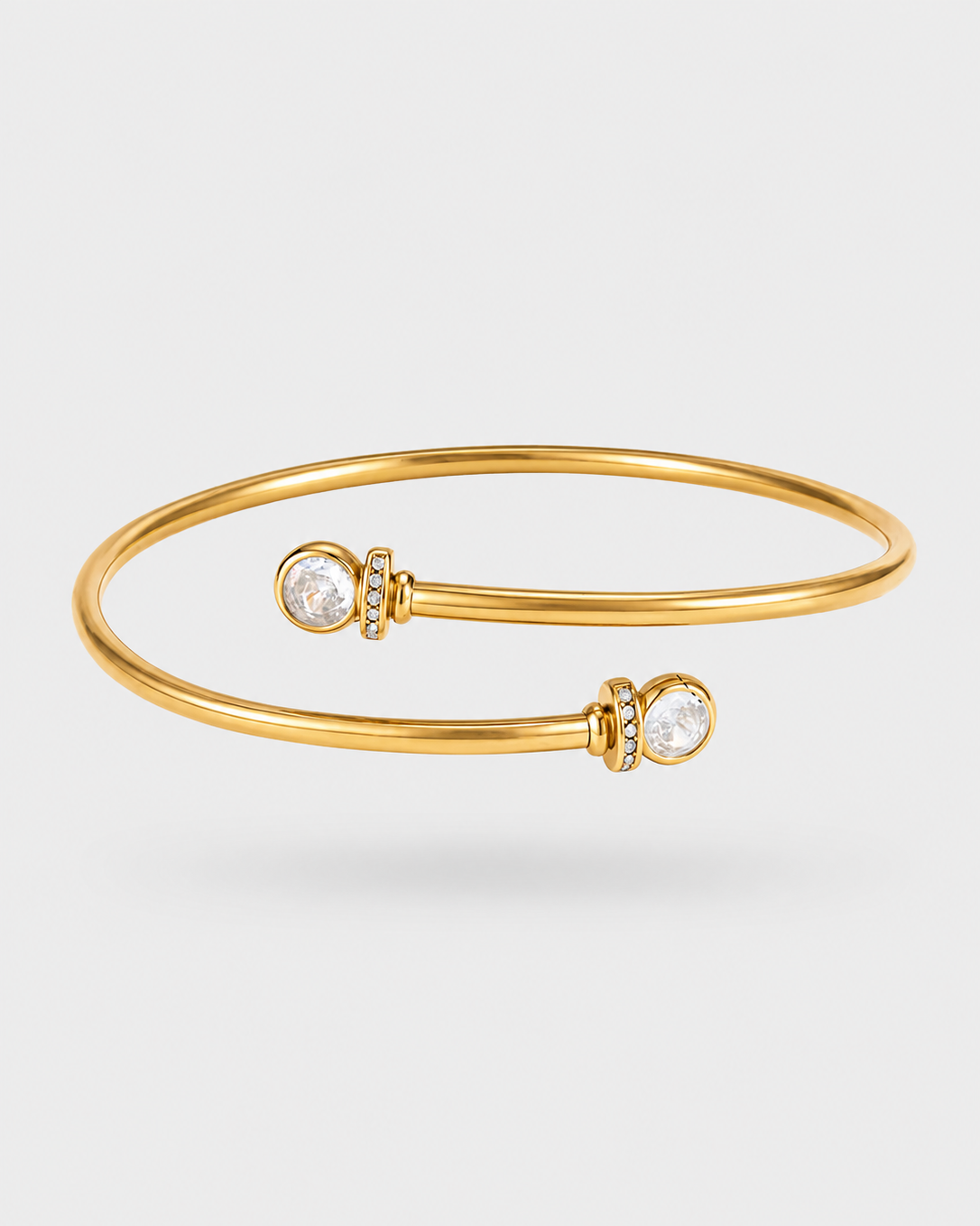 A gold bangle bracelet with two clear round bezel-set stones, displayed on a light gray background.