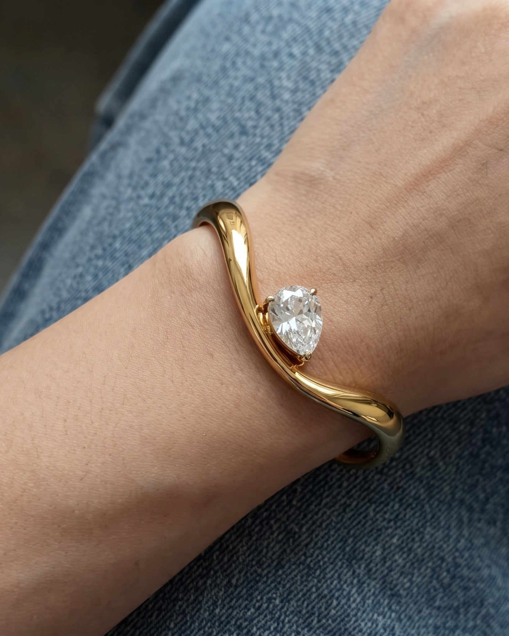 A gold cuff bracelet with a heart-shaped clear gemstone, worn on a person's wrist over blue denim jeans.