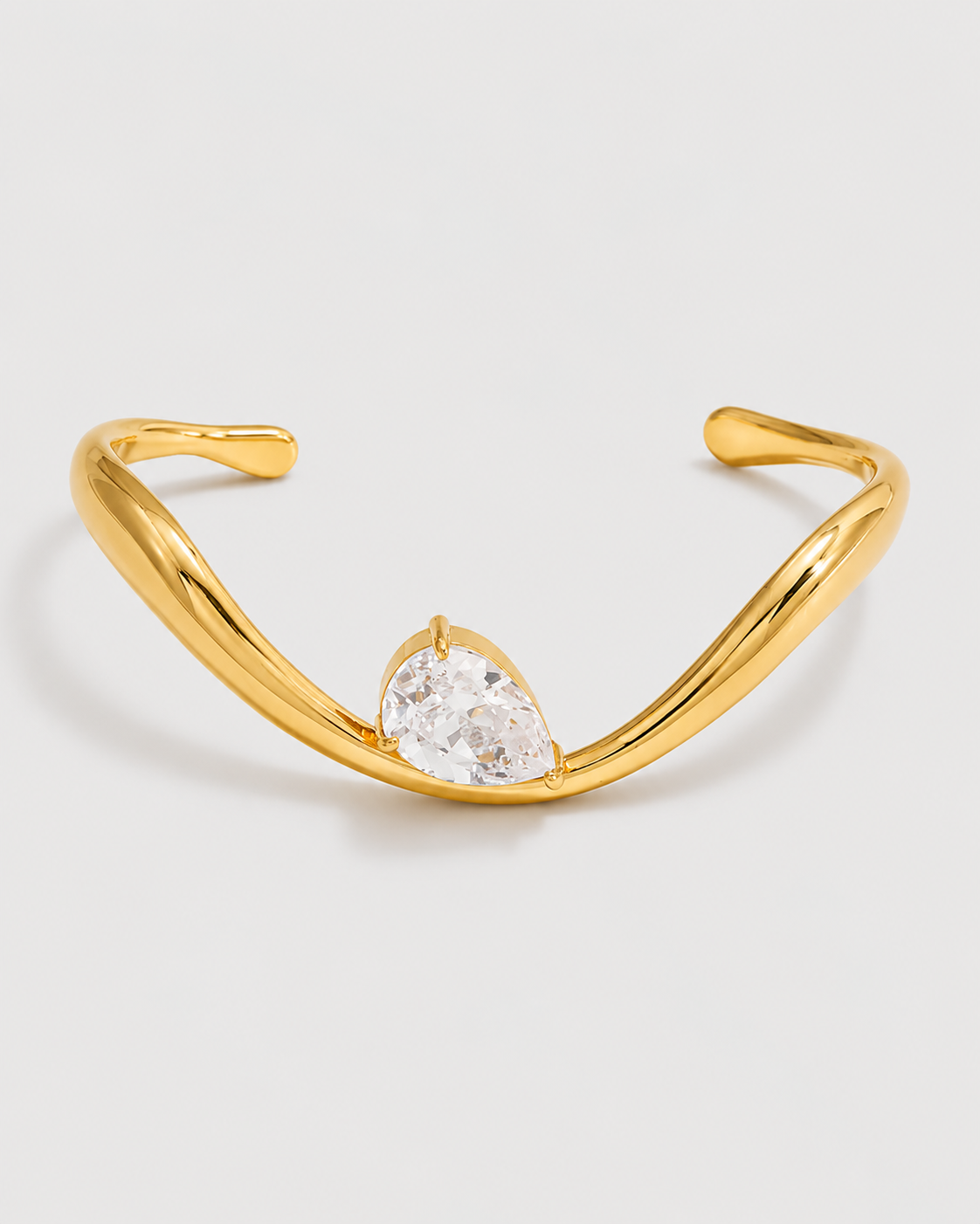 A polished gold-toned cuff bracelet featuring a pear-cut clear gemstone at the center, set against a clean light gray background.