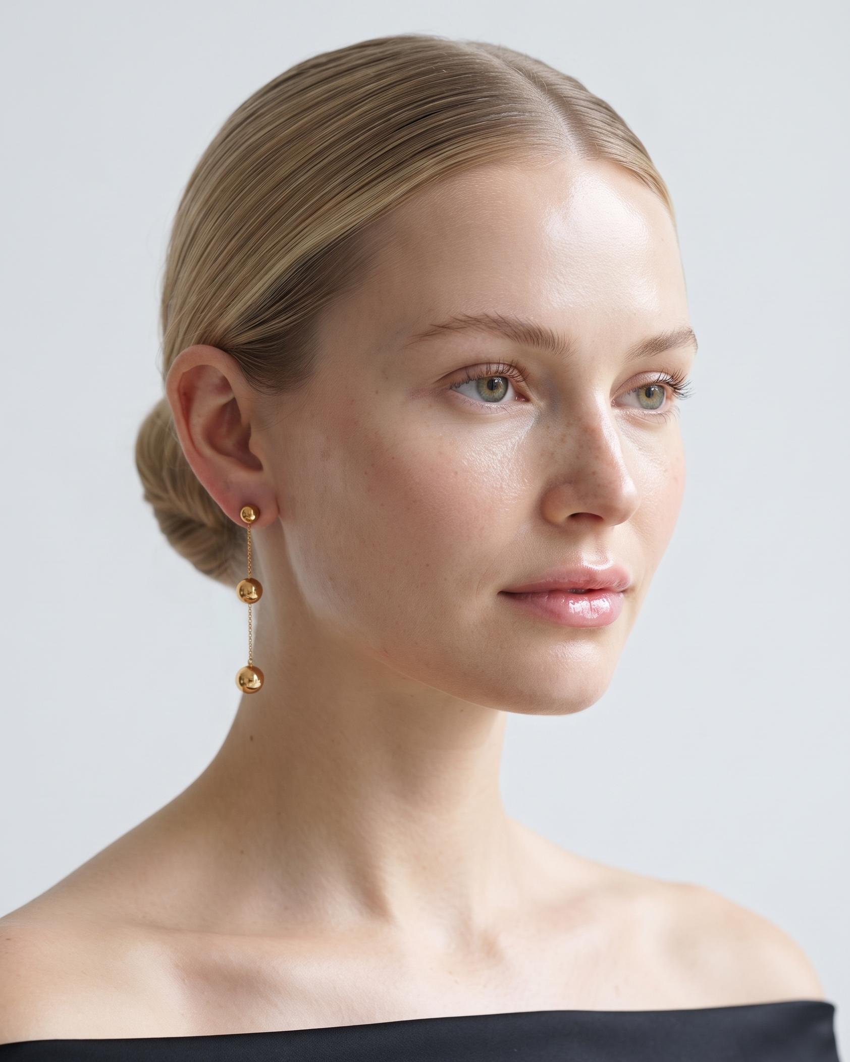A close-up portrait of a woman with blonde hair pulled back, wearing a delicate gold dangle earring with three stacked spherical beads. She is wearing an off-the-shoulder black top against a clean, light gray studio background.