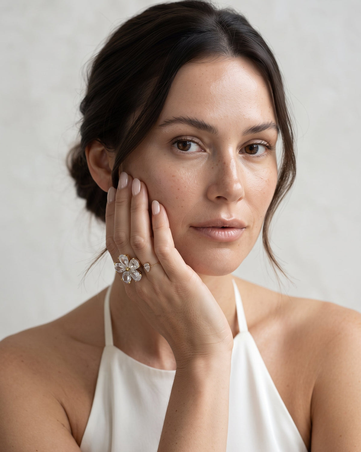 A close-up portrait of a woman with dark hair in a low bun, wearing a white halter top and a large silver flower-shaped ring with clear stones, touching her cheek against a light textured background.