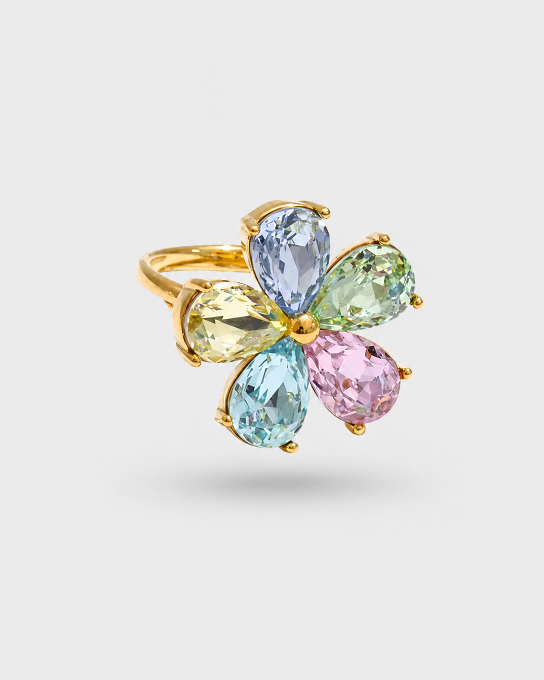 A gold flower-shaped ring featuring five teardrop-cut gemstones in pastel shades of blue, green, yellow, pink, and light aqua, set against a light gray background.