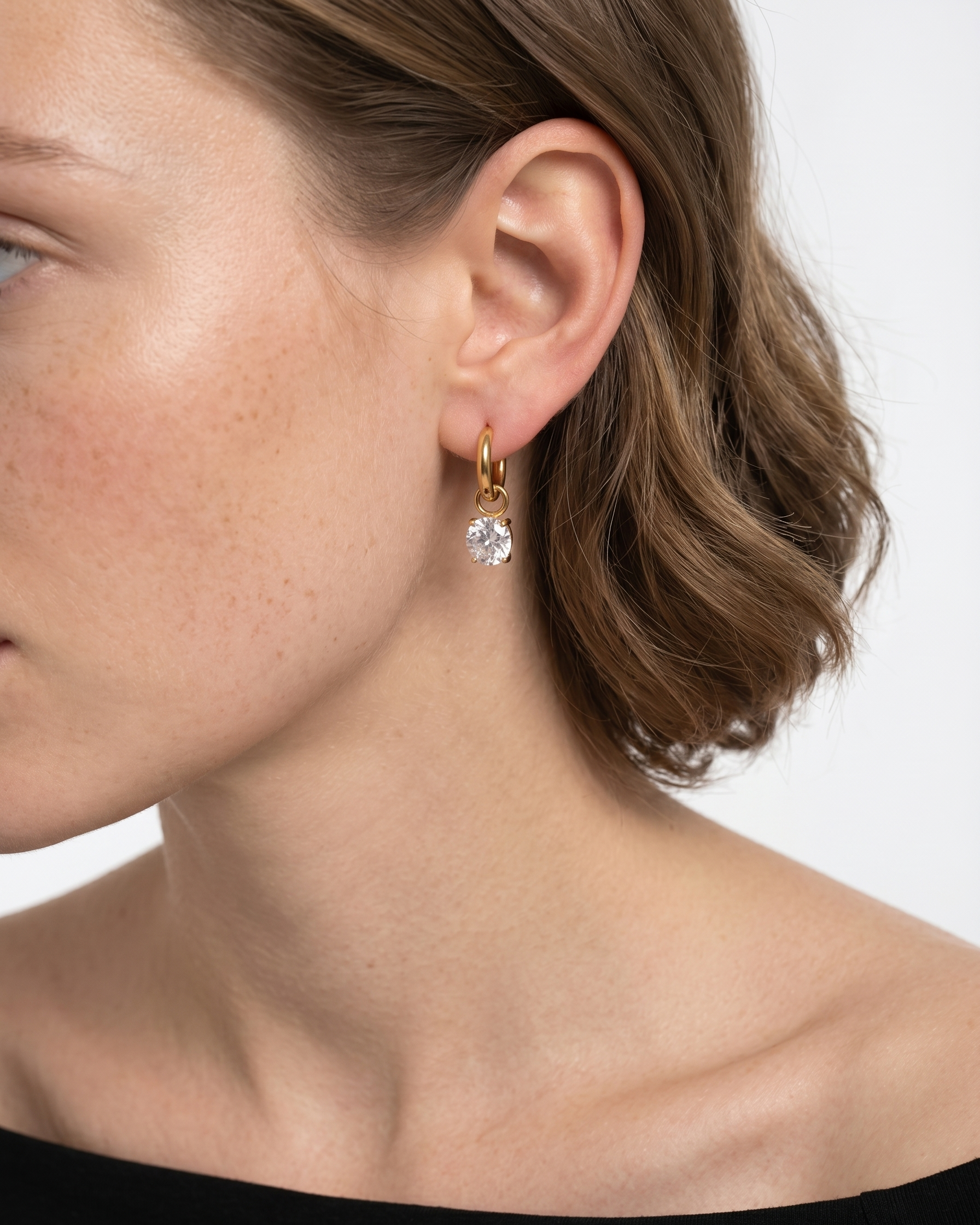 Close-up of a person wearing a gold earring with a clear gemstone on a neutral background