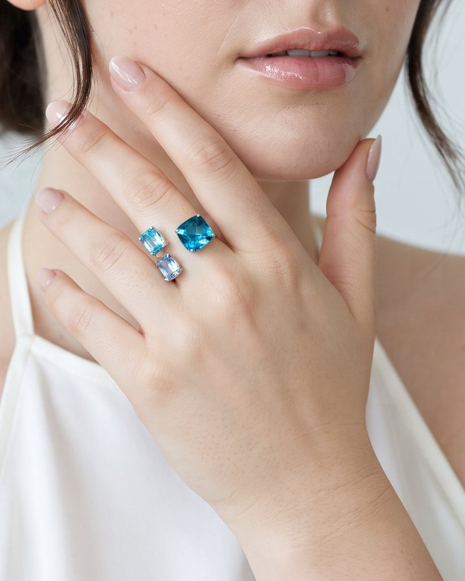 A woman with dark hair in an updo wearing a white halter top, showcasing a silver ring with two blue rectangular gemstones on her finger, set against a light gray background.