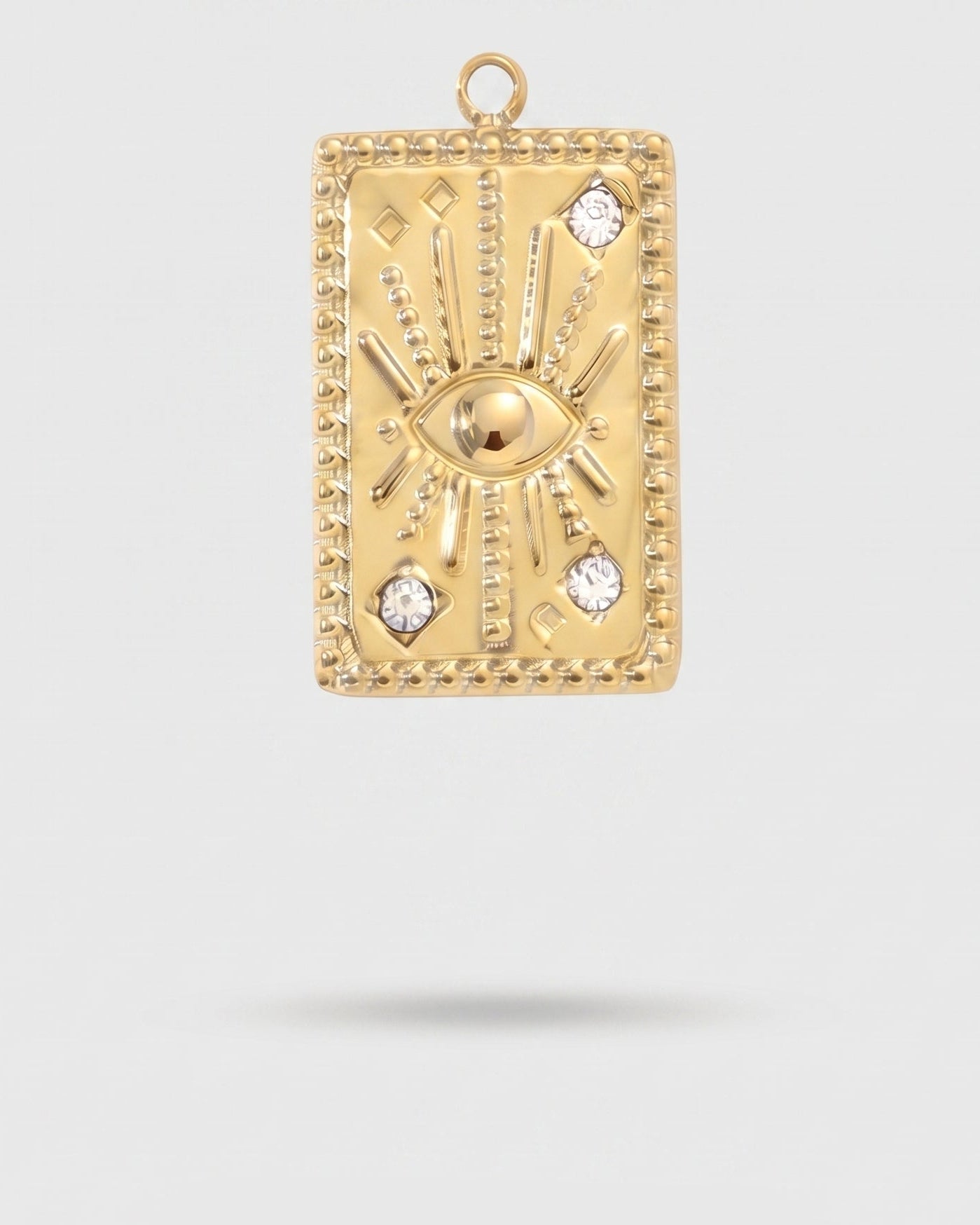 A gold rectangular pendant featuring a central eye motif with radiating sunburst lines and small clear stones, shown against a light gray background with a soft shadow below.