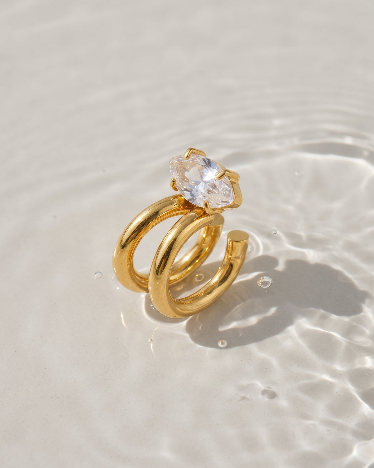 A gold ring with a clear heart-shaped gemstone, resting on a light beige sandy surface with shallow water ripples and reflections.