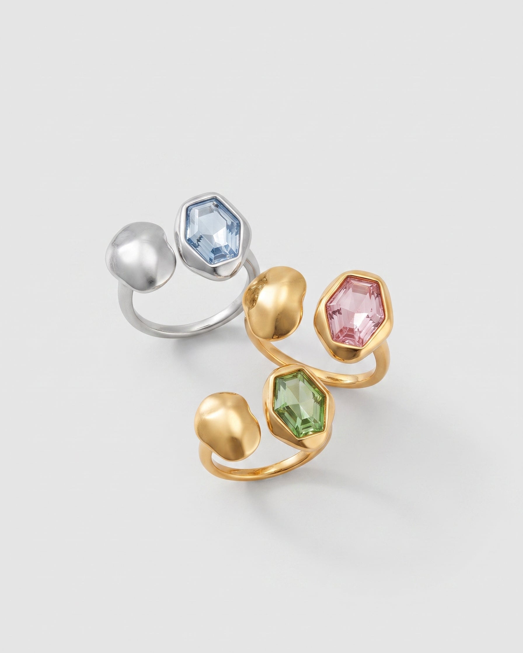Two sculptural open rings on a light gray background. One ring is silver with a light blue hexagonal gemstone, and the other is gold with pink, green, and blue hexagonal gemstones.
