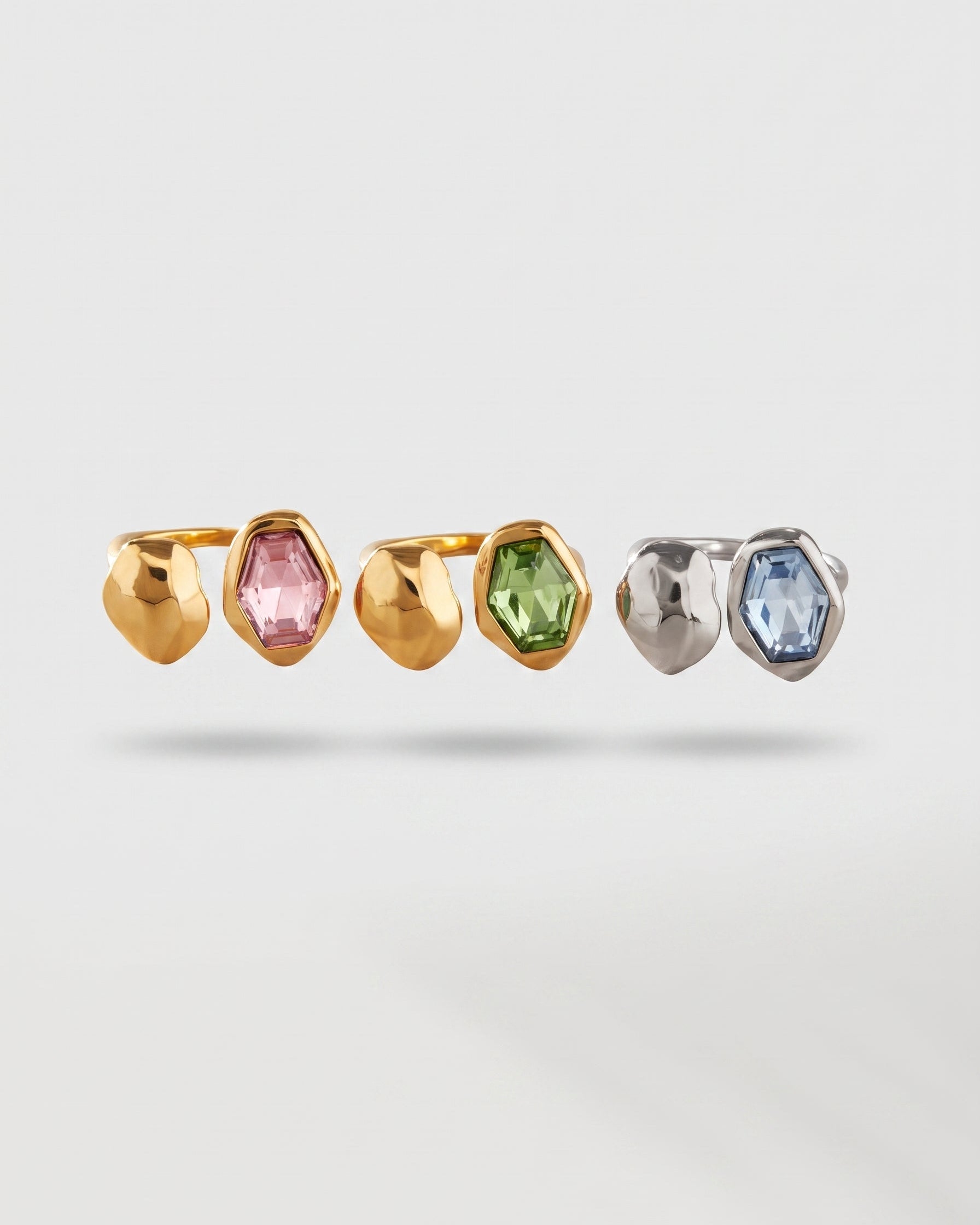 Three small geometric stud earrings arranged in a row on a light gray background. Each earring has a faceted metal setting with a gemstone: two are gold-toned with pink and green stones, and one is silver-toned with a light blue stone.