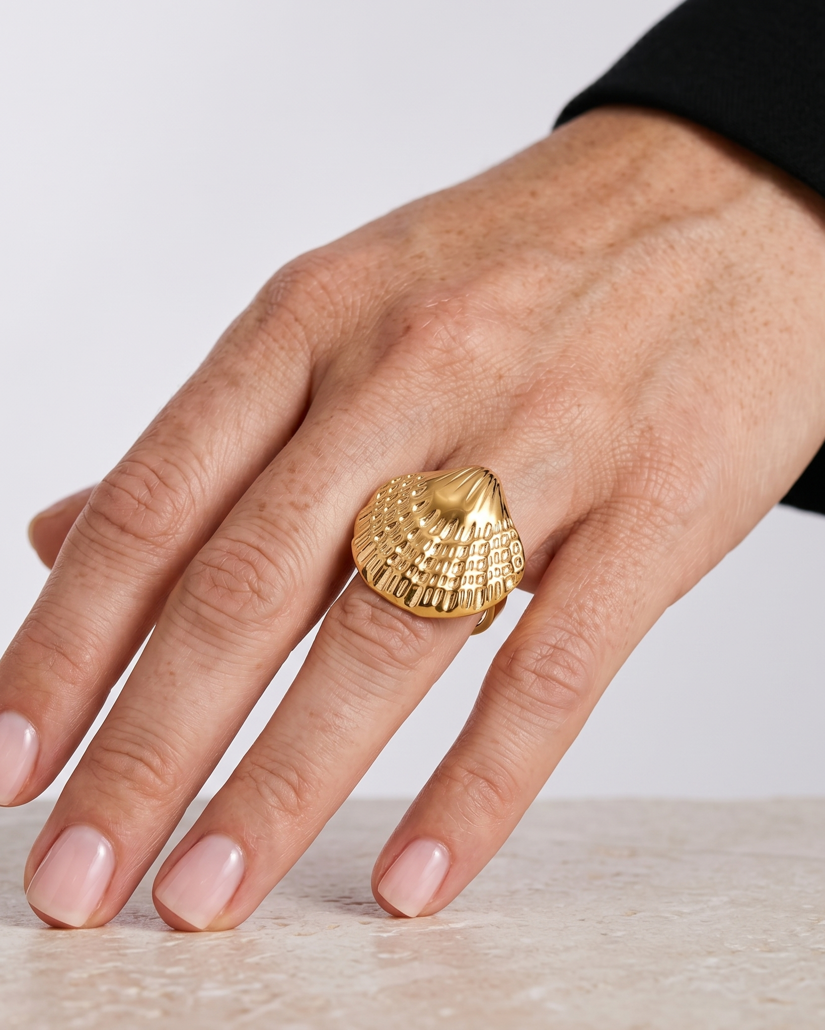 Gold shell-shaped ring on a hand against a neutral background