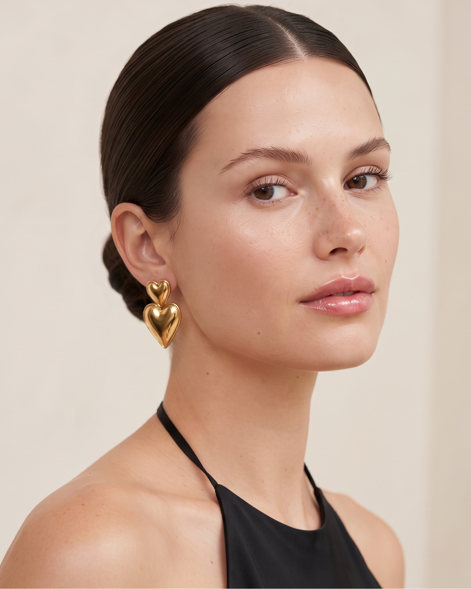 A close-up portrait of a woman with dark hair pulled back, wearing a shiny gold heart-shaped stud earring and a black halter top against a soft beige background.
