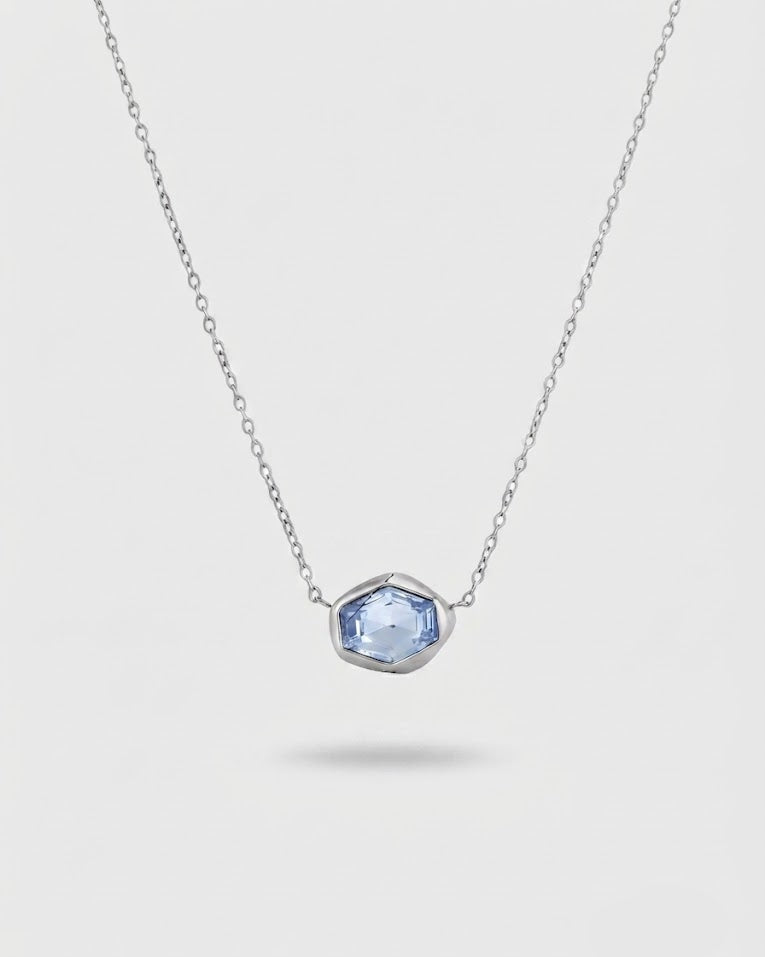 A delicate silver chain necklace featuring a small, irregular-cut light blue gemstone pendant in a silver bezel setting, shown against a light gray background.