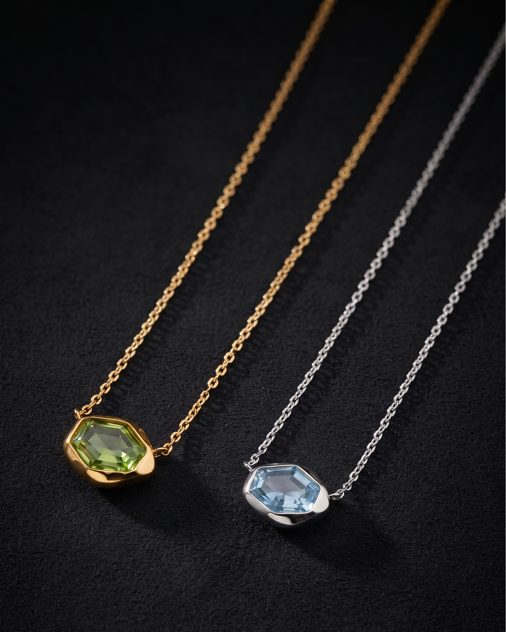 Two delicate necklaces with faceted gemstone pendants are displayed on a dark textured background. One necklace is gold with a green gemstone, and the other is silver with a light blue gemstone.