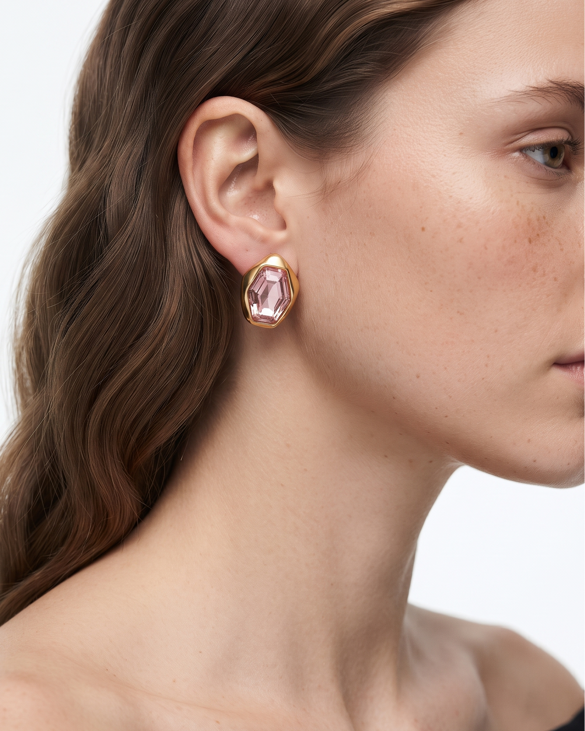 A close-up side profile of a woman wearing a gold-toned drop earring with a faceted pink gemstone, set against a clean white background.