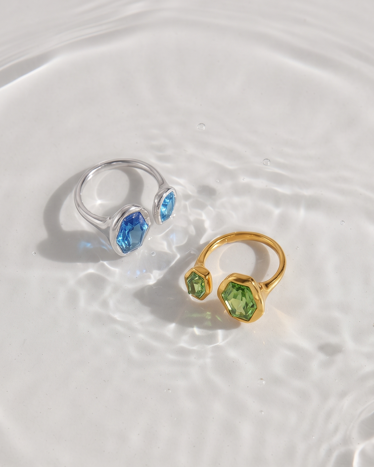 Two open ring necklaces with marquise-cut gemstones, one in silver with blue stones and one in gold with green stones, resting on a rippled white water surface.